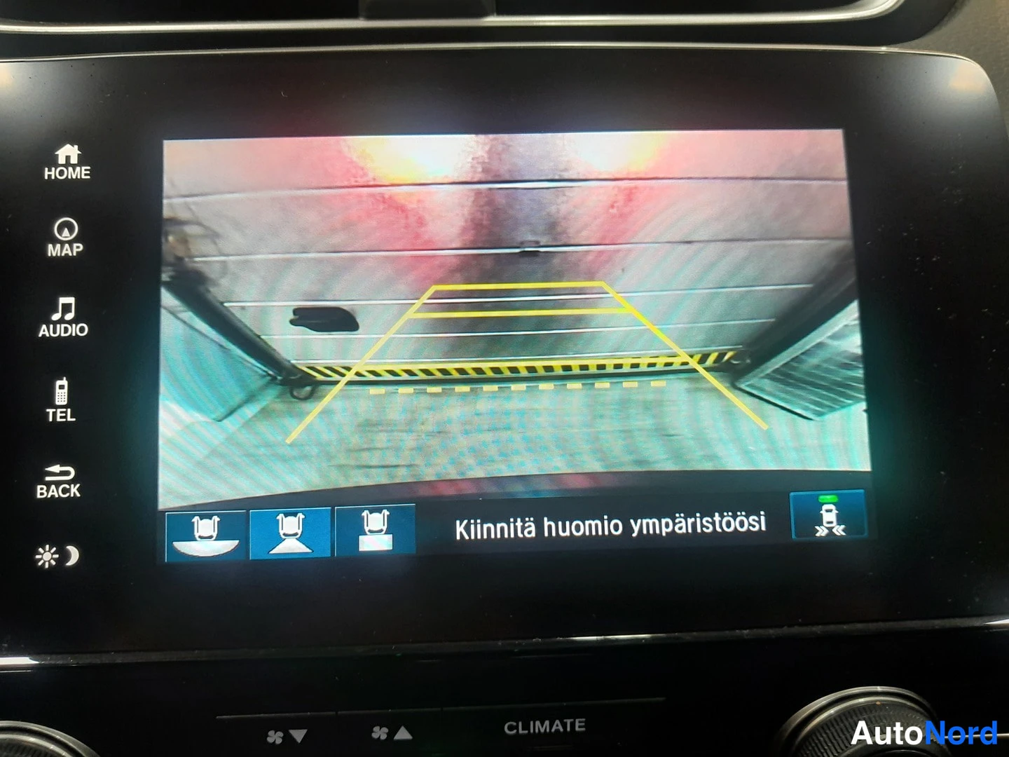 Yksityiskohta: Auton näyttö näyttää taaksepäin katsovan kuvan varoituslinjineen ja tekstillä Kiinnitä huomion ympäristöösi sekä ISOFIX-merkkejä