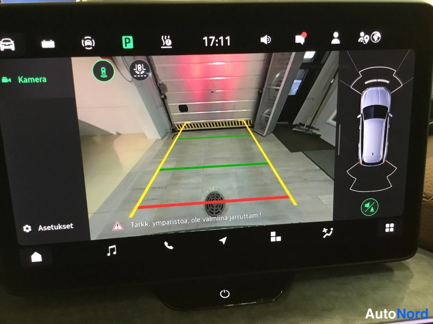 Takaa: Auton infotainment-näyttö taaksepäin käännetyllä kameranäkymällä, ympäristövaroituksella 'Tarkk. ympäristöä, ole valmiina jarruttama!' ja ajoneuvon ylhäältä kuv