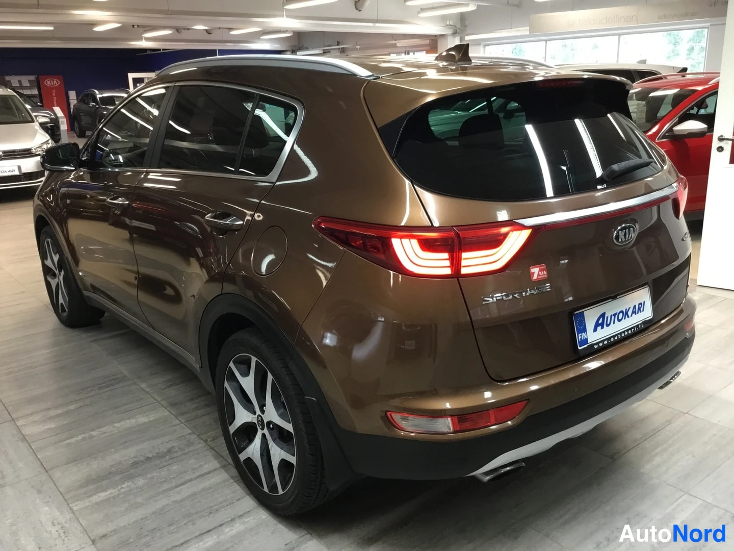 Takaviisto: Kia Sportage pronssinruskea takakulmasta vasemmalta, takavalot päällä, mallimerkintä SPORTAGE konepellissä.