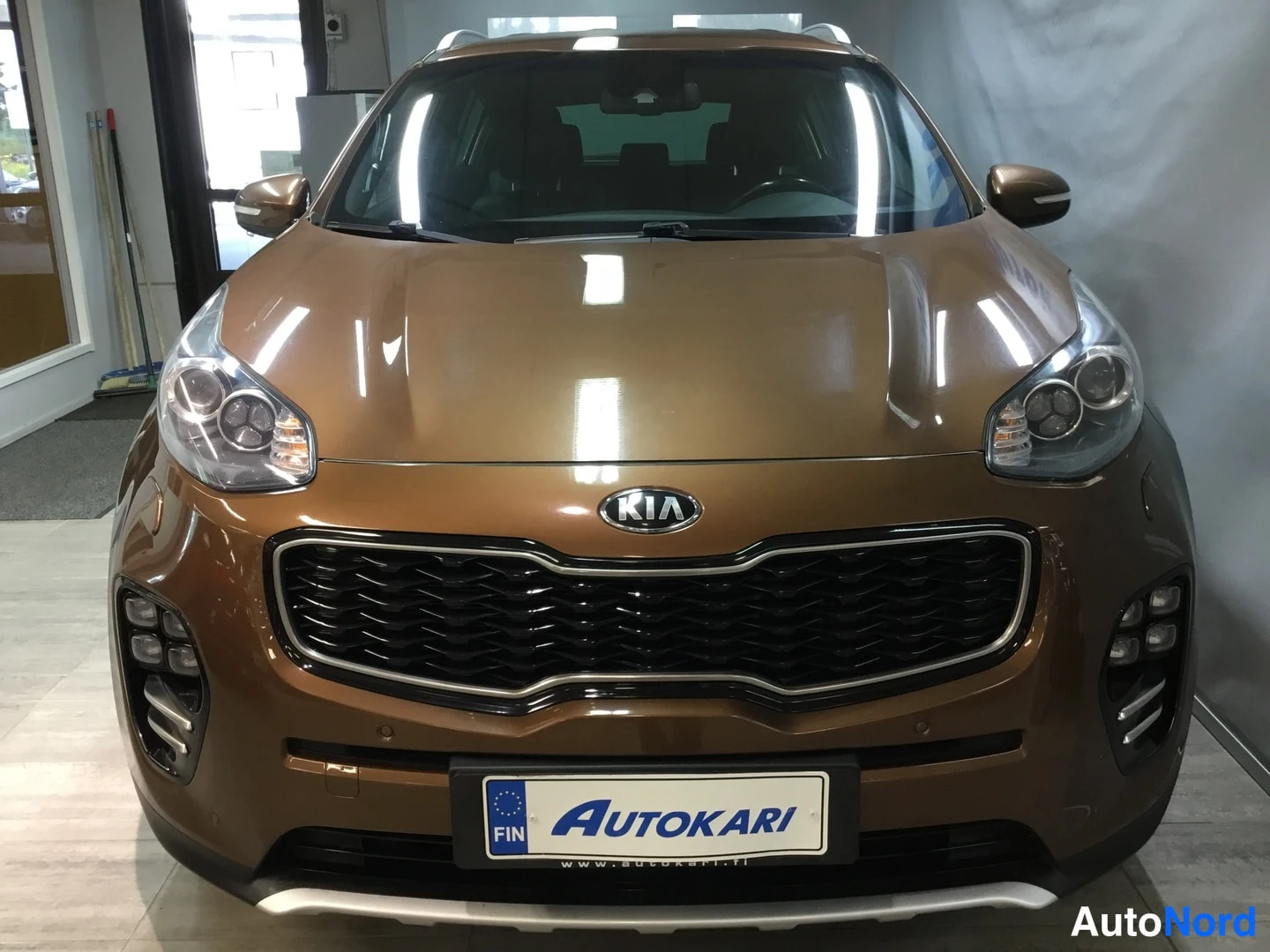 Etuviisto, nokka oikealle: Front three-quarter view of brown SUV, showing grille with KIA badge and right-side headlight assembly.