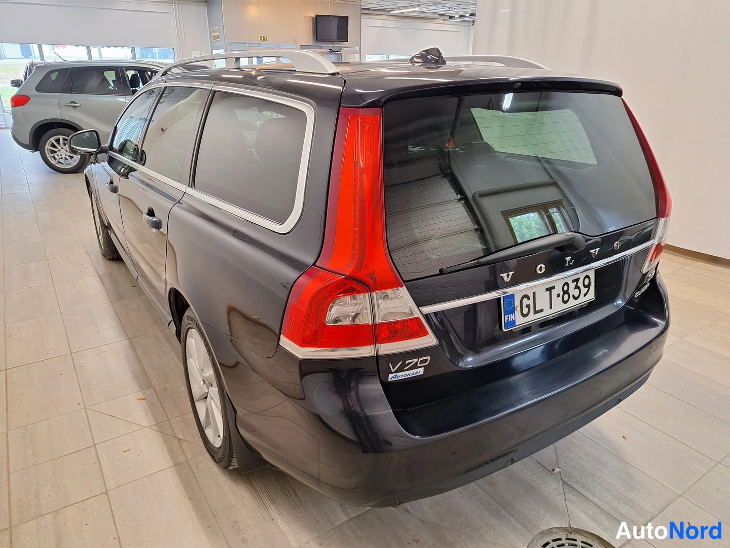 Takaviisto: Tumma farmariauto takaa sivulta kuvattuna, V70-merkintä ja rekisterikilpi näkyvissä.