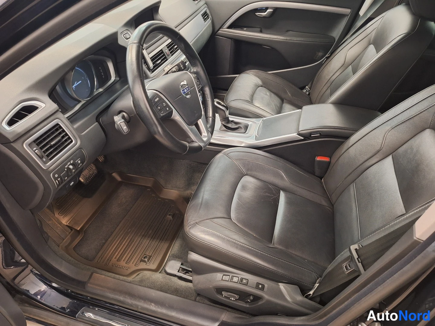 Muu: Volvo V70:n sisätilat kuljettajan puolelta: ohjauspyörä, center console, mustat nahkaistuimet ja kellertävä askelmatto.