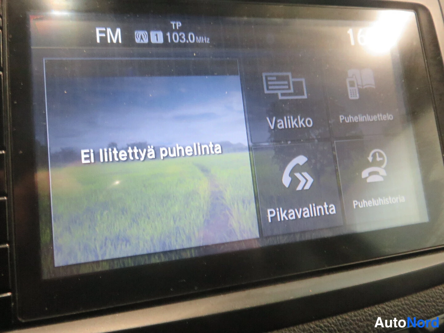 Yksityiskohta: Auton näyttö radiovastaanotolla FM 103.0 MHz ja puhelinvalikko, jossa teksti 'Ei liitettyä puhelinta' sekä valikot Valikko, Puhelinluettelo, Pikavalinta ja Puhe