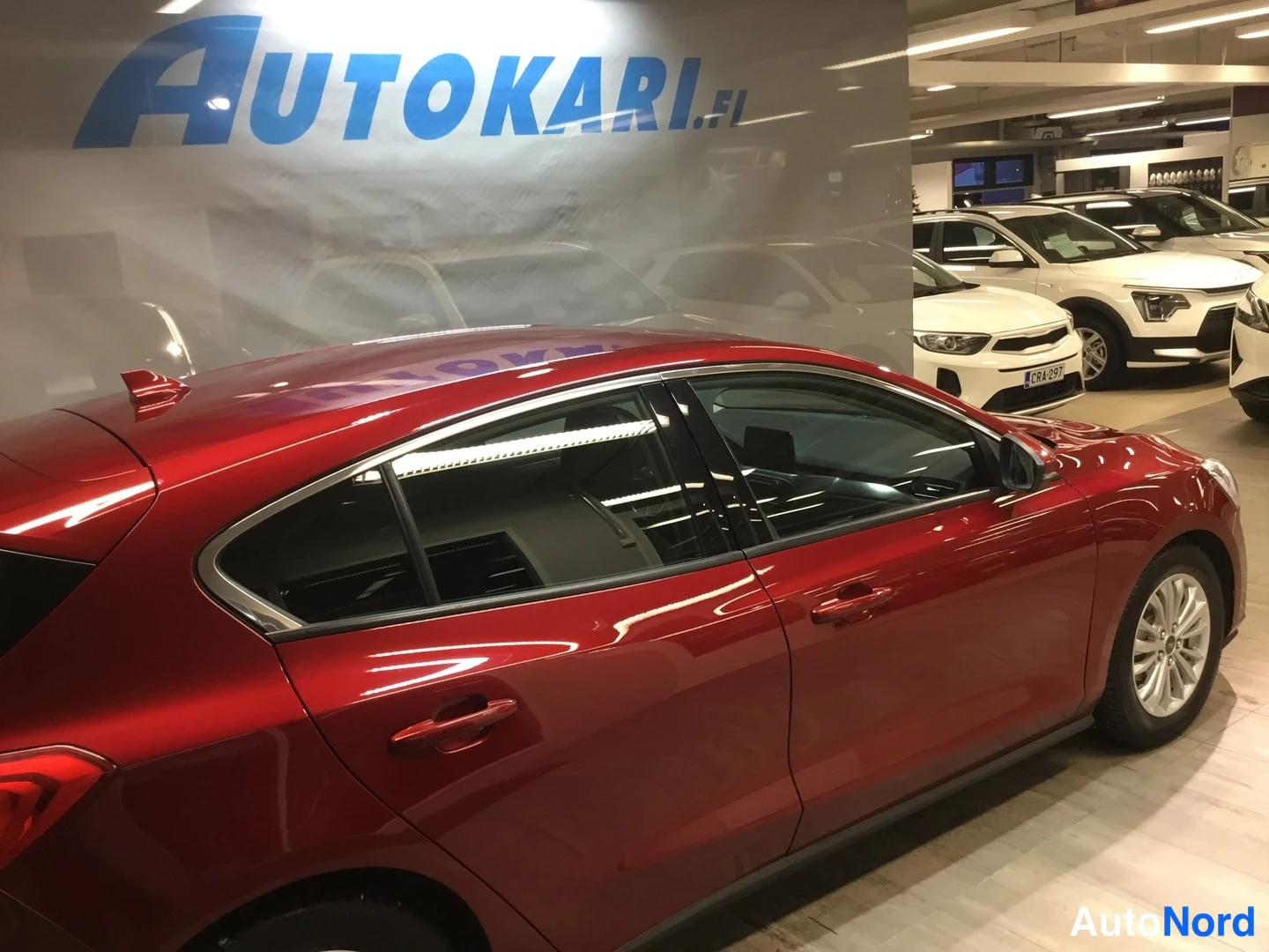 Takaviisto: Punaisen Ford Focusin takaosa viistosti vasemmalta autosalissa; sivutakovalo ja kylki näkyvät taustalla muita autoja.