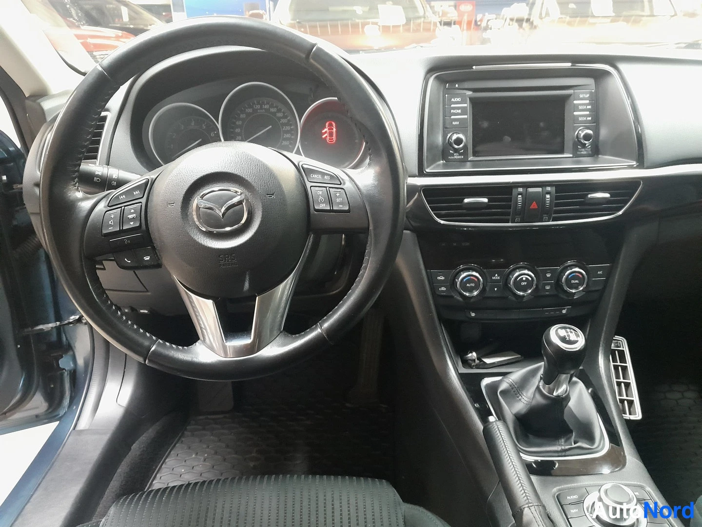 Edestä: Mazdan ohjaamo vasemmalta ohjattuna: Mazda-ohjauspyörä, manuaalivaihteisto, kojetaulun mittarit ja infotainment-näyttö.