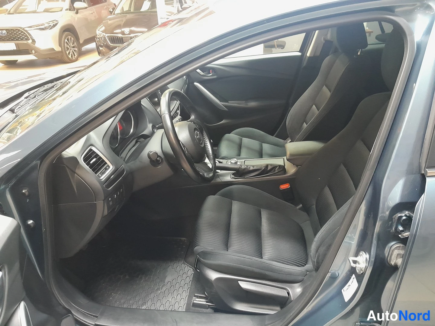 Edestä: Kuljettajan ovi auki: Mazda 6:n etummat istuimet ja ohjaamo tummilla kankaistuilla istuimilla.