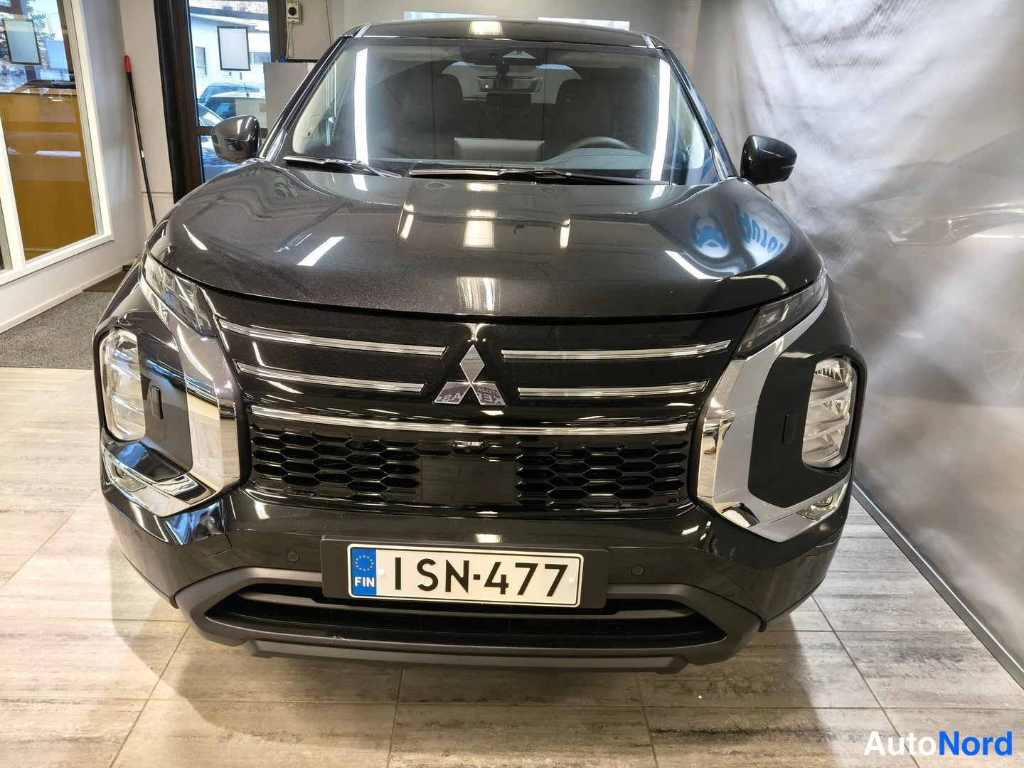 Edestä: Sisävalokuva musta Mitsubishi Outlander -henkilöauto suoraan edestäpäin, Mitsubishi-tähti etusäleikössä ja rekisterikilpi.