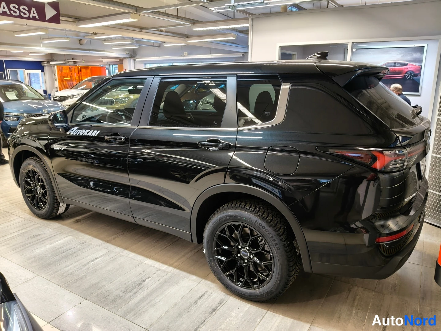 Takaviisto: Autokari-näyttelytilassa sisävalokuva musta Mitsubishi Outlander -henkilöauto takaa vasemmalta kolmannesnäkymä, mustat vanteet ja Autokari-teksti kuljettajan or
