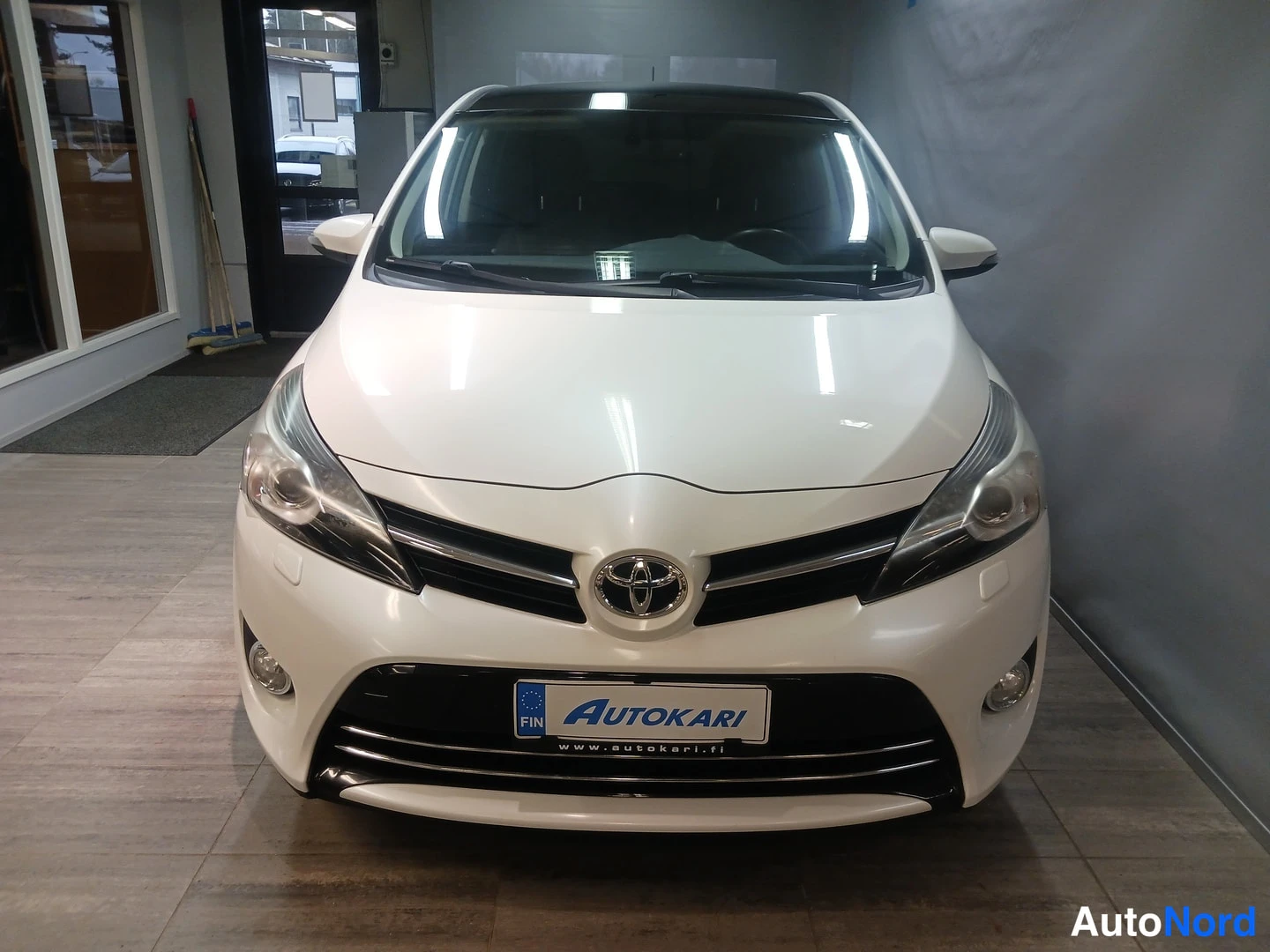 Edestä: Auton etupää suoraan edestä kuvattuna: valkoinen Toyota-merkki, etumoottoritila ja Autokari-tunnus rekisterikilven paikalla sisätiloissa.