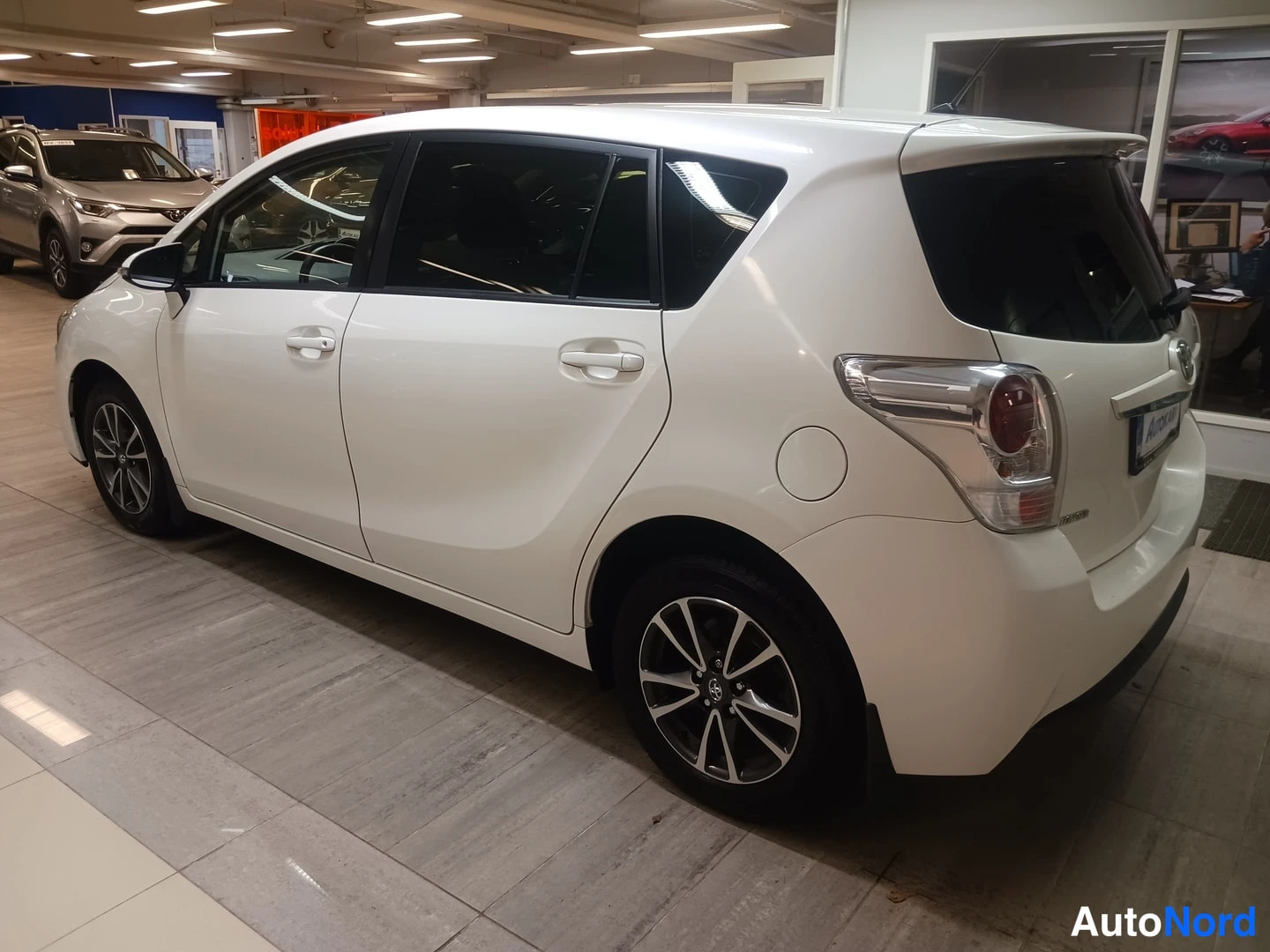 Takaviisto: Valkoinen Toyota Verso takakulmasta vasemmalle suunnattuna: takapuskuri, takavalot ja kattospoileri näkyvät sisätiloissa.