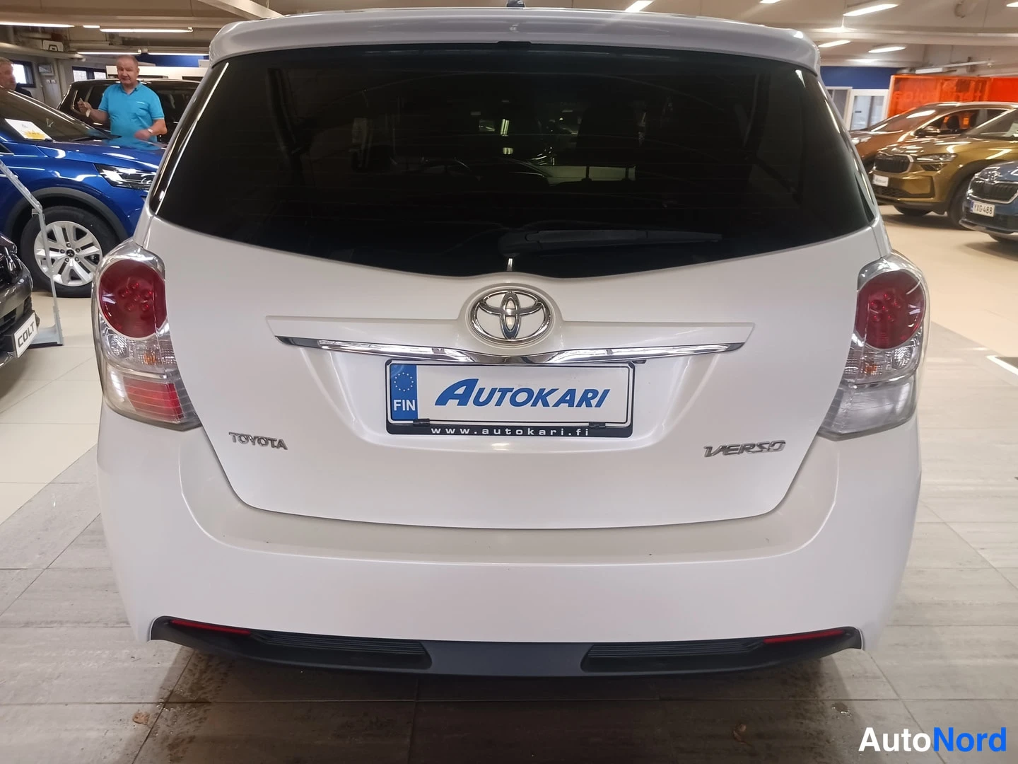Takaa: Auton takapää suoraan takaa kuvattuna: valkoinen Toyota Verso -merkki, takalasinpyyhkijä ja Autokari-tunnus rekisterikilven paikalla sisätiloissa.