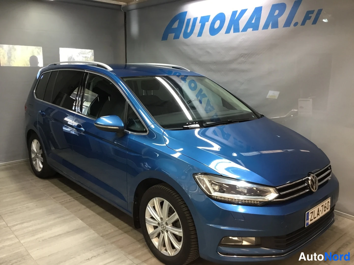 Etuviisto, nokka oikealle: Sininen Volkswagen Touran etukulmasta oikealta, nokka osoittaa kuvan oikeaan reunaan, rekisterikilpi.