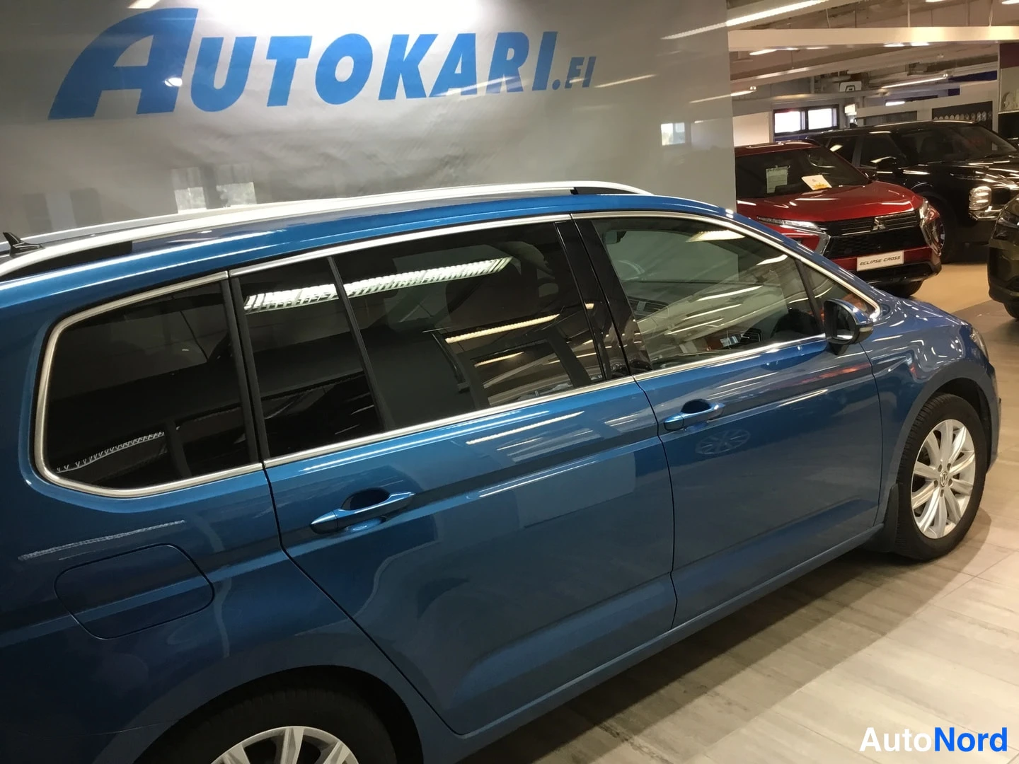 Sivulta: Sinisen Volkswagen Touranin sivuprofiili näkyvissä sisätiloissa, kylki ja takapään pyörä näkyvät.