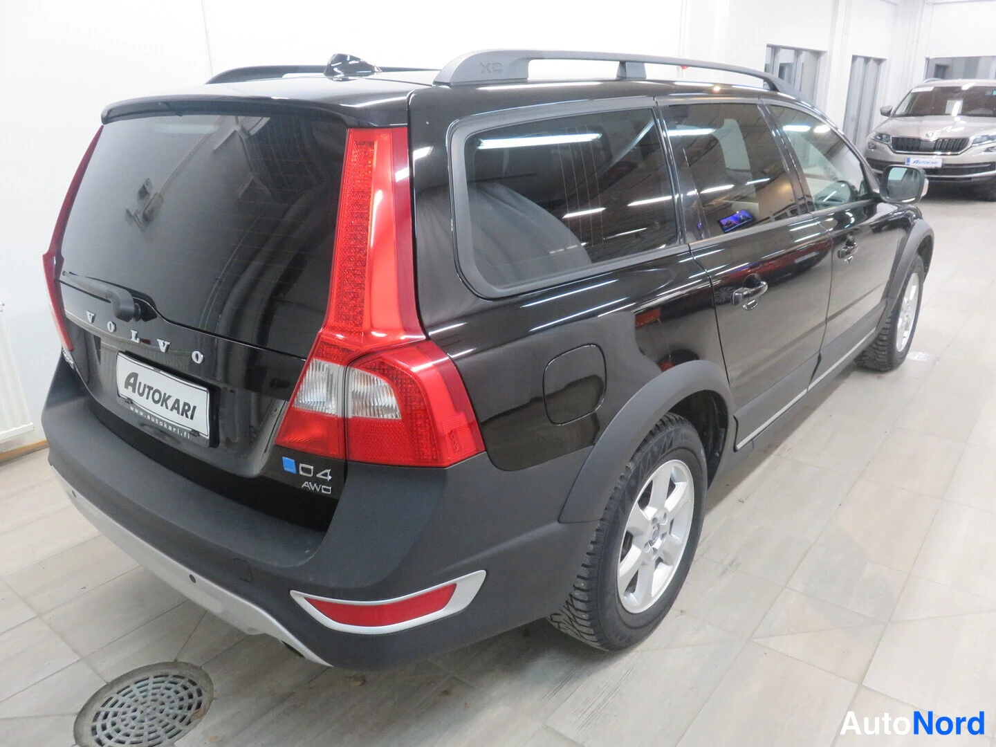 Takaviisto: Mustan farmariauton taka- ja kylkinäkymä sisätiloissa; perässä VOLVO- ja D4 AWD -tekstit sekä AUTOKARI-kyltti, XC-kattokaiteet.