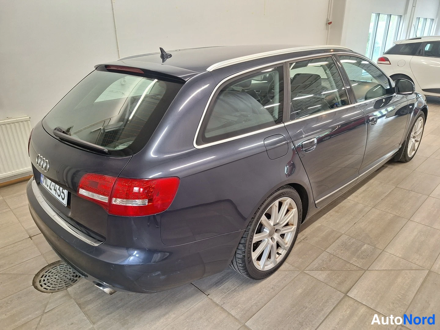 Takaviisto: Rear three-quarter view of a dark blue Audi A6 Avant from the passenger side, showing the chrome window trim, silver roof rails, alloy wheels, and red tail ligh