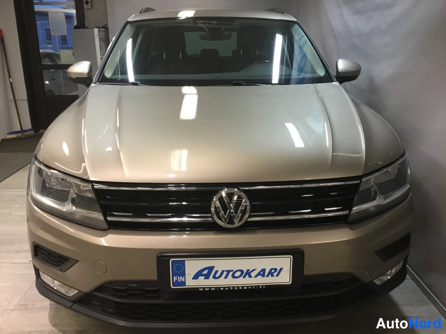 Edestä: Volkswagen Tiguinin etupää näkyy suoraan edestä: etusäleikkö VW-merkillä, ohjaamovalot ja etupuskuri ovat näkyvissä, koko auto on beige-kultainen.