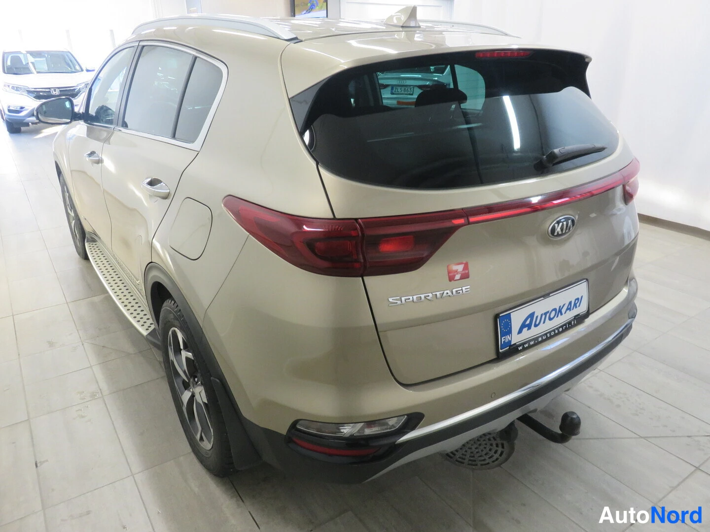 Takaviisto: Beige Kia Sportage takaa oikealta kulmasta kuvattuna autotallissa; punaiset takavalot, Sportage-tunnus ja Autokari-rekisterikilven suojus näkyvät.