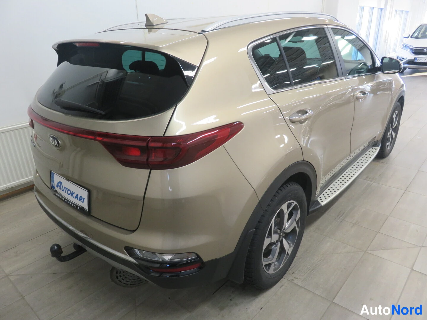 Takaviisto: Beige Kia Sportage takaa vasemmalta kulmasta kuvattuna autotallissa; paneelikatto, vetokoukku ja auton kylki näkyvät.