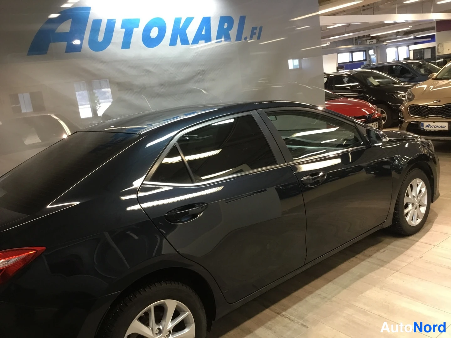 Takaviisto: Tummansininen sedan auto takaviistöstä katsottuna auton myyntitilassa, taustalla muita autoja.