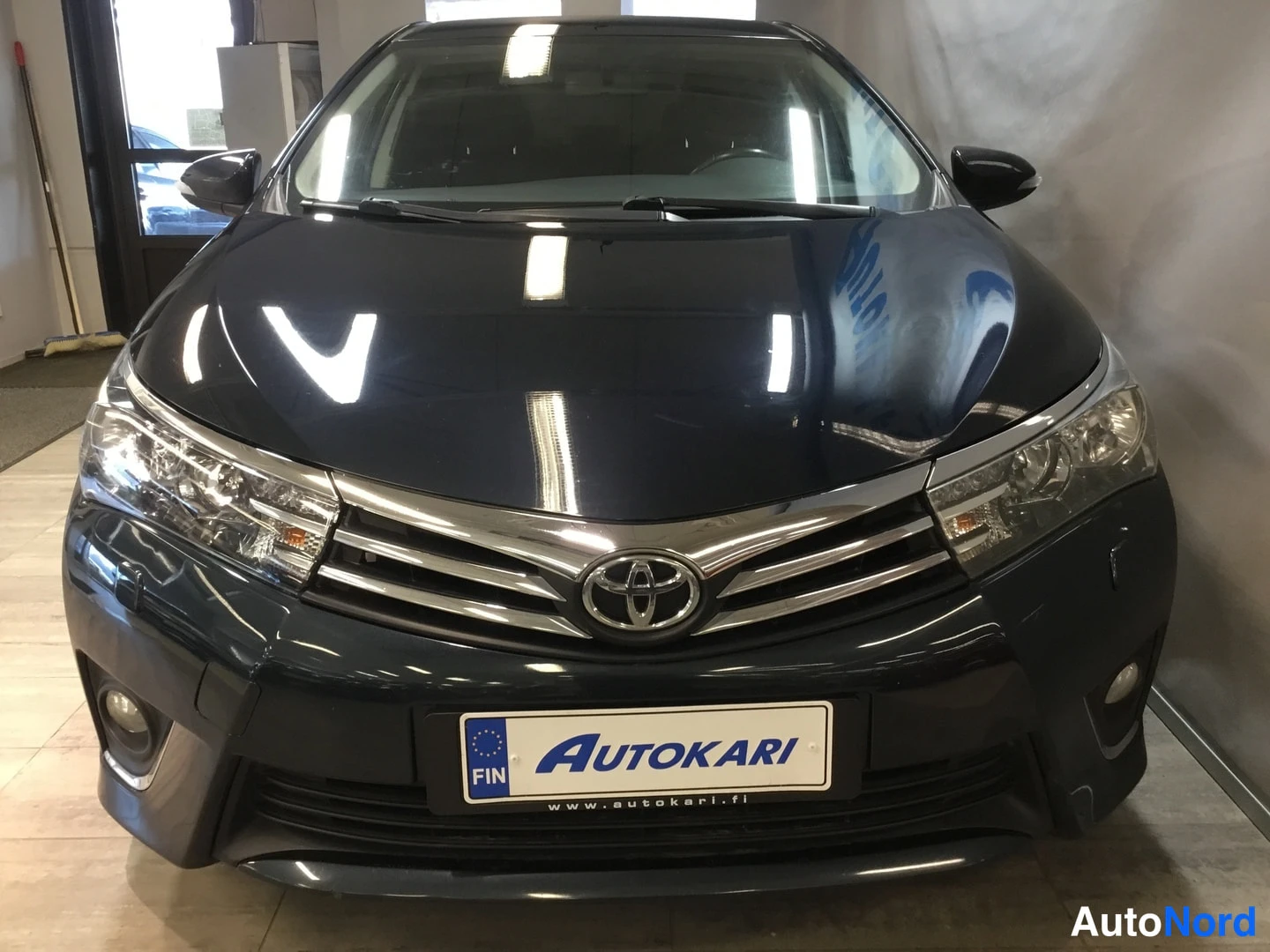 Edestä: Mustan Toyota Corollan etupää edestäpäin, Toyota-merkki keskellä etusäleikköä, Autokari-kyltti rekisterin paikalla.