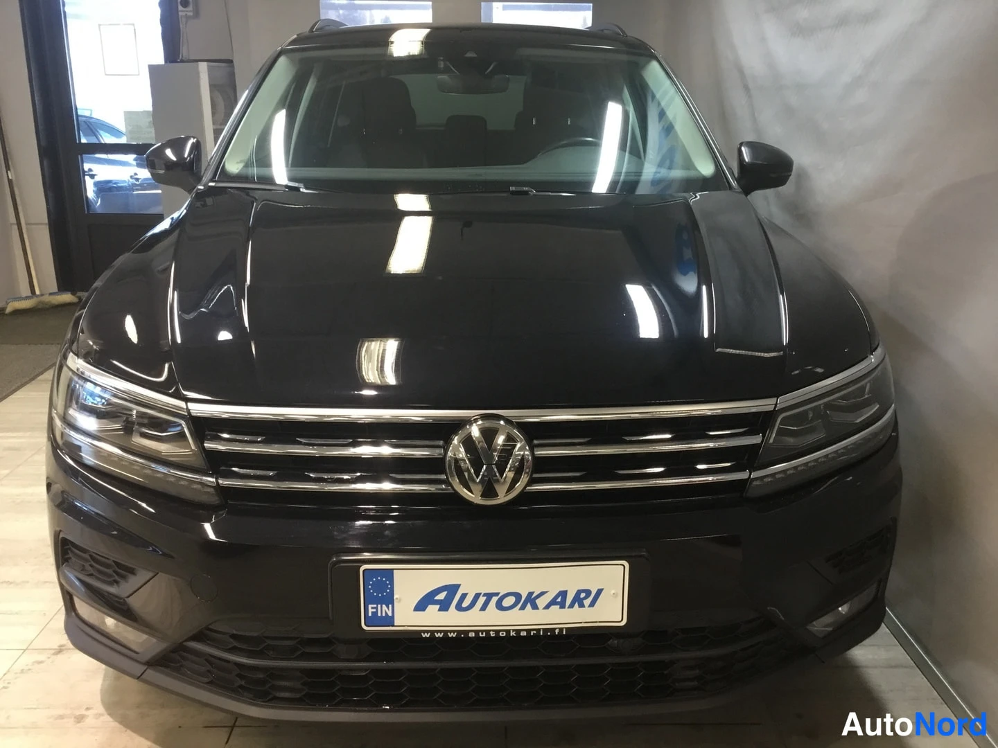 Edestä: Mustan Volkswagen Tiguanin etupää kohtisuoraan edestä, VW-merkki jäähdyttimeen ja Autokari-rekisterikilpi.