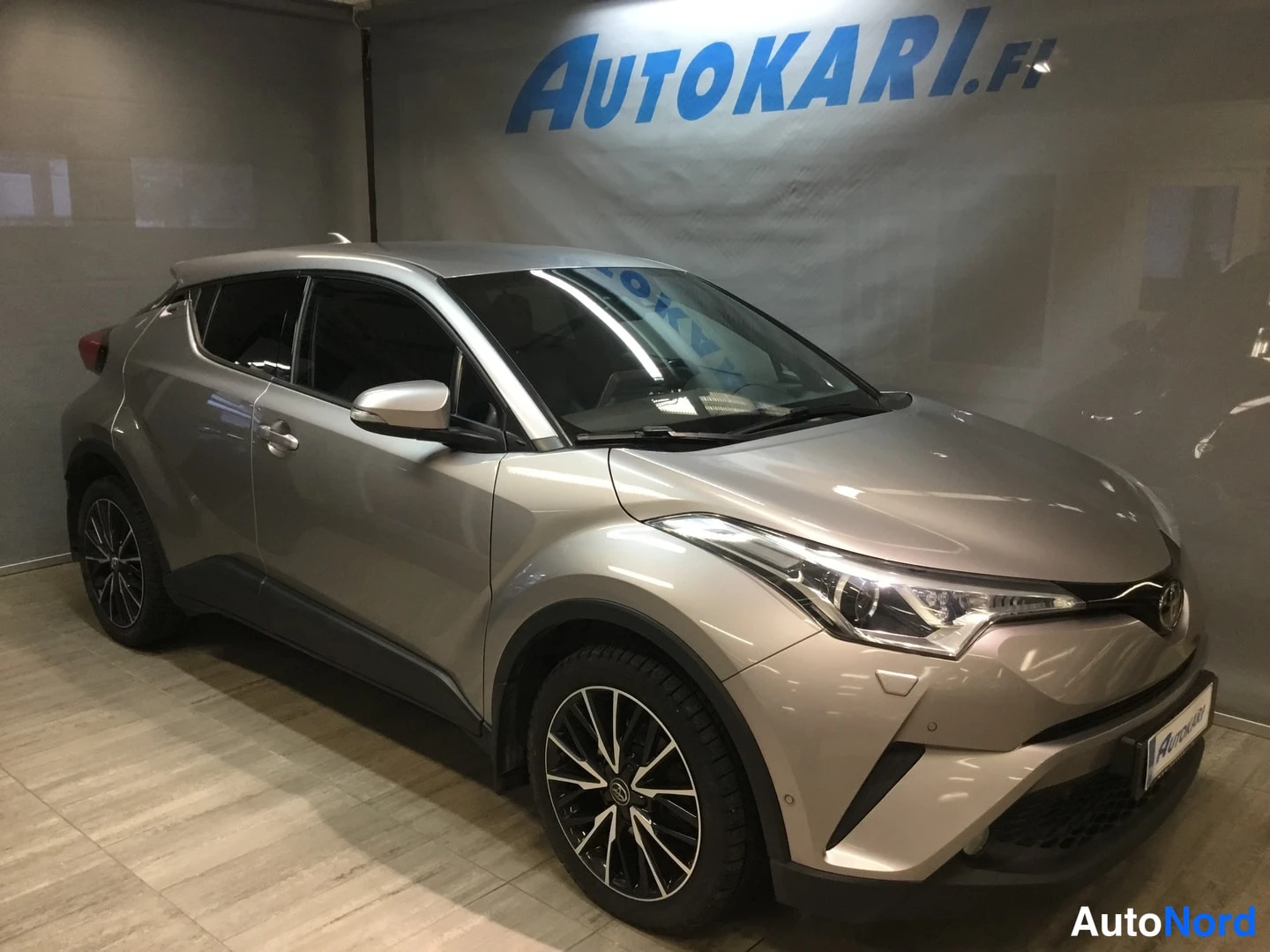 Etuviisto, nokka oikealle: Toyota C-HR hopeanvärisenä kulmassa etu-OV, keulakuva oikealle, myymälätilassa Autokari.fi-taustapressin edessä.