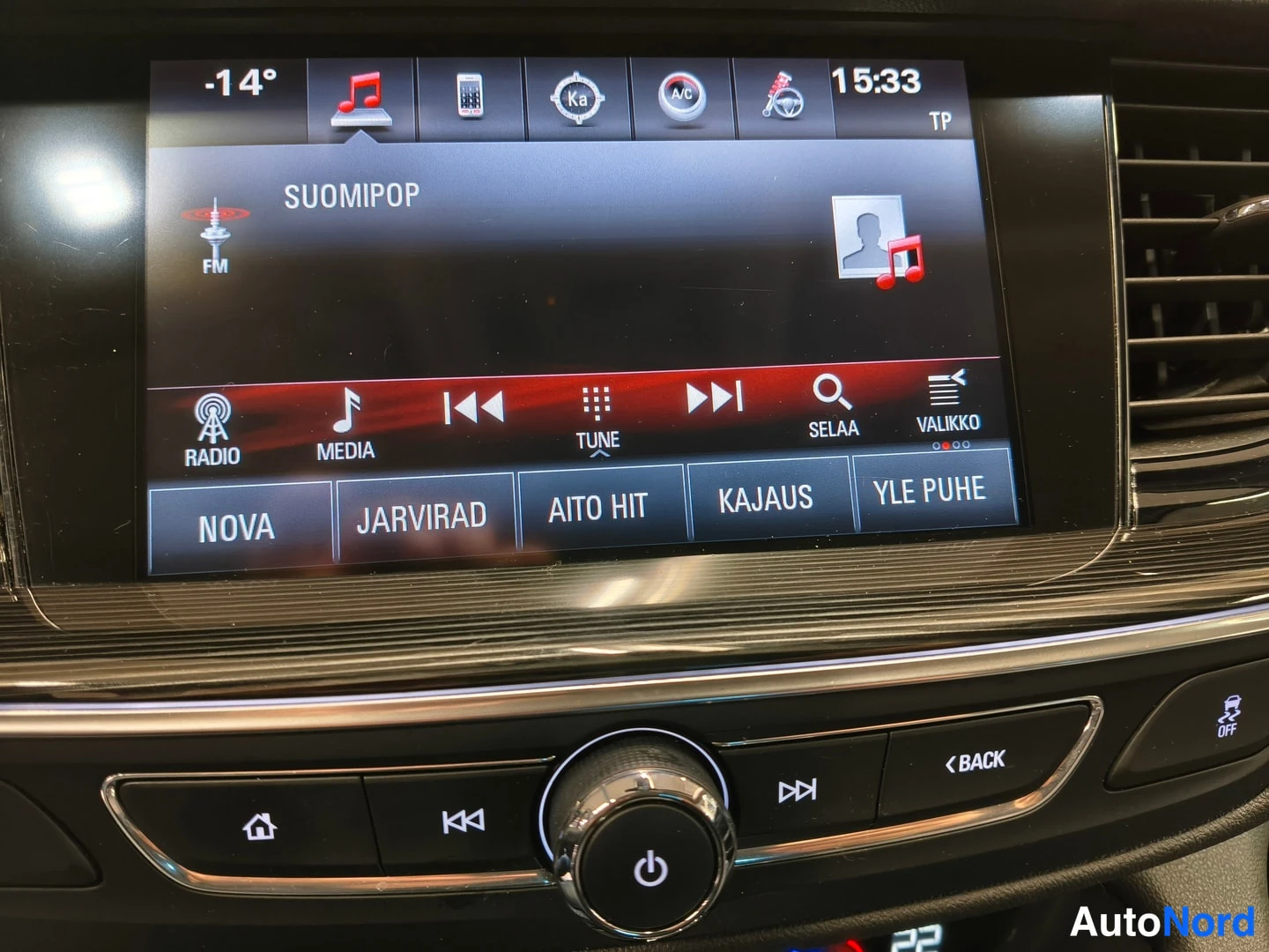 Yksityiskohta: Auton infotainment-näyttö radiotoiminnossa, FM-kanava SUOMIPOP ja esiasetukset NOVA, JARVIRAD, AITO HIT, KAJAUS, YLE PUHE; alla multimediasäädimet ja BACK-nappi