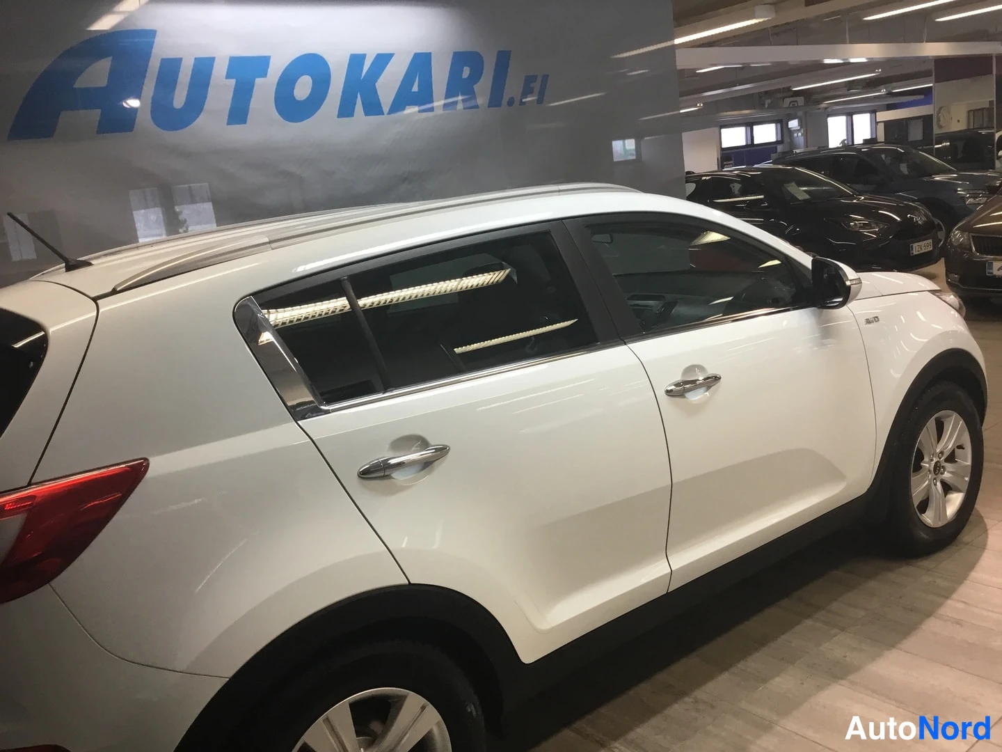 Sivulta: Valkoinen henkilöauto sivusta kuvattuna, takapuskuri kuvan vasemmassa reunassa ja etupyörä oikealla.