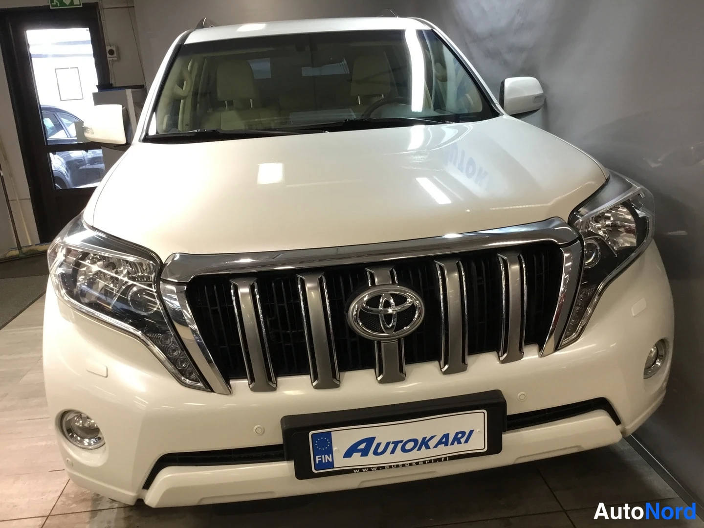 Edestä: Toyota Land Cruiser, etukulma. Valkoinen koripinta-ala, kromattu etusäleikkö Toyota-emblemillä, sumuvalot ja Autokari-tunnus etupuskurissa.