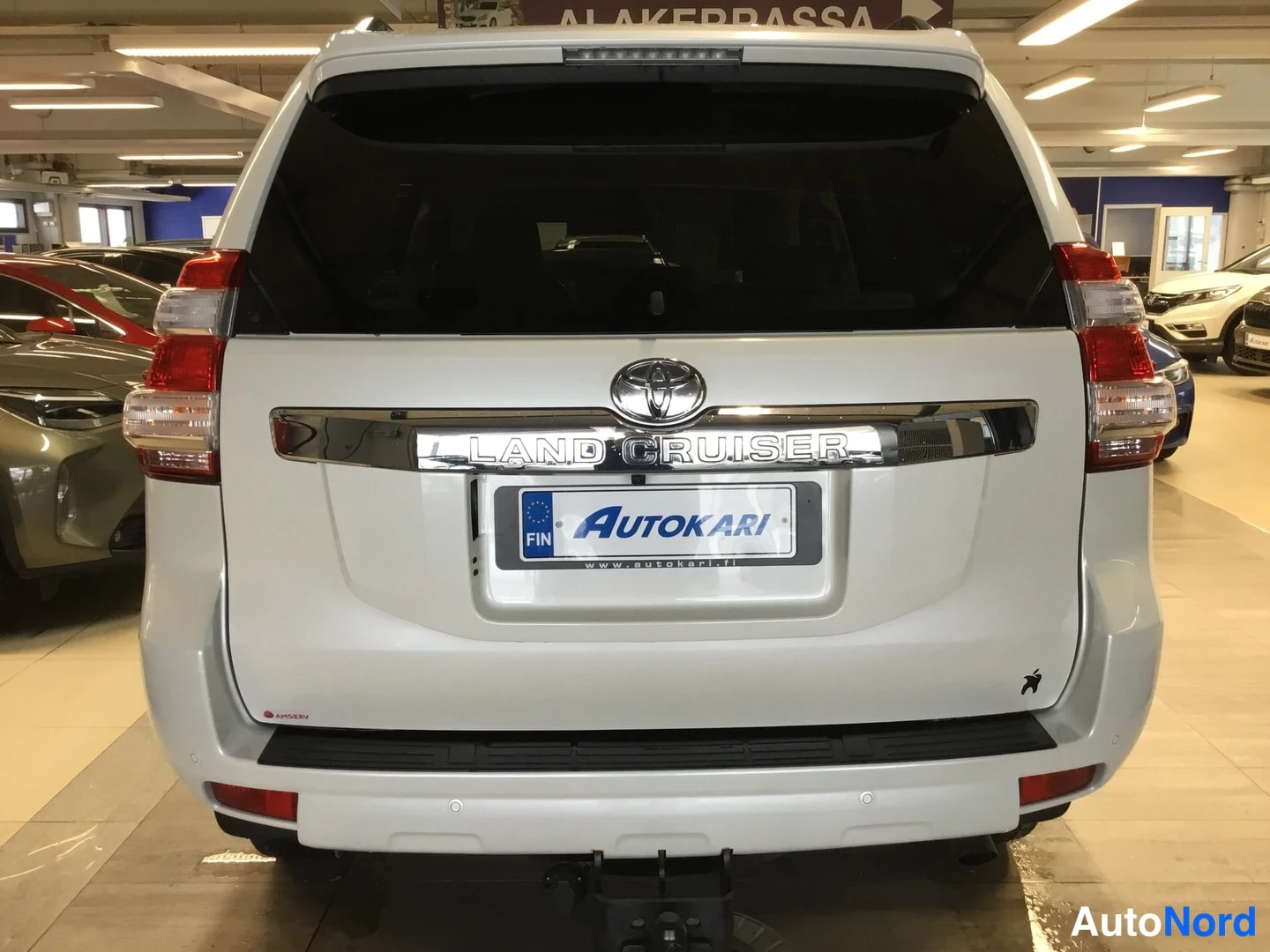 Takaa: Toyota Land Cruiser, takakulma. Valkoinen koripinta-ala, LAND CRUISER -teksti takaluukussa, Autokari-tunnus, ajoneuvonperäkkäisjärjestys ja vetokoukku kuvassa.