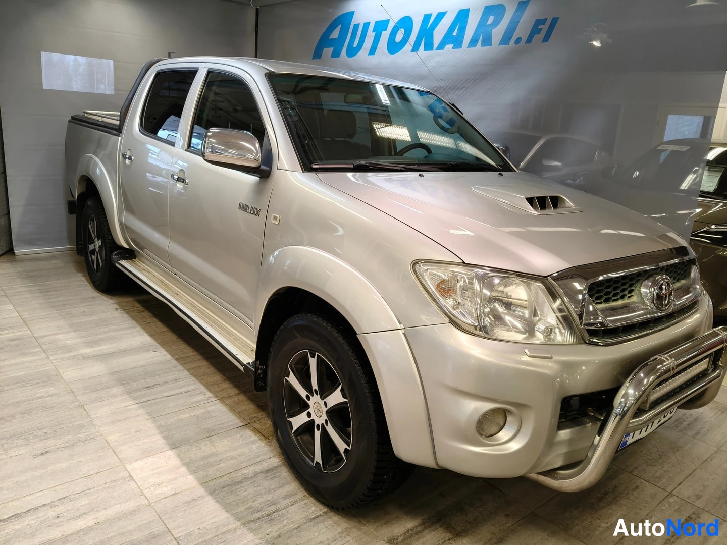 Etuviisto, nokka oikealle: Hopea Toyota Hilux -mallinen pakettiauto nähtynä kolmannes­kulmasta keulasta oikealle, kromikeulakaari etupuskurissa ja sivupeilat kyljissä, esittelytila