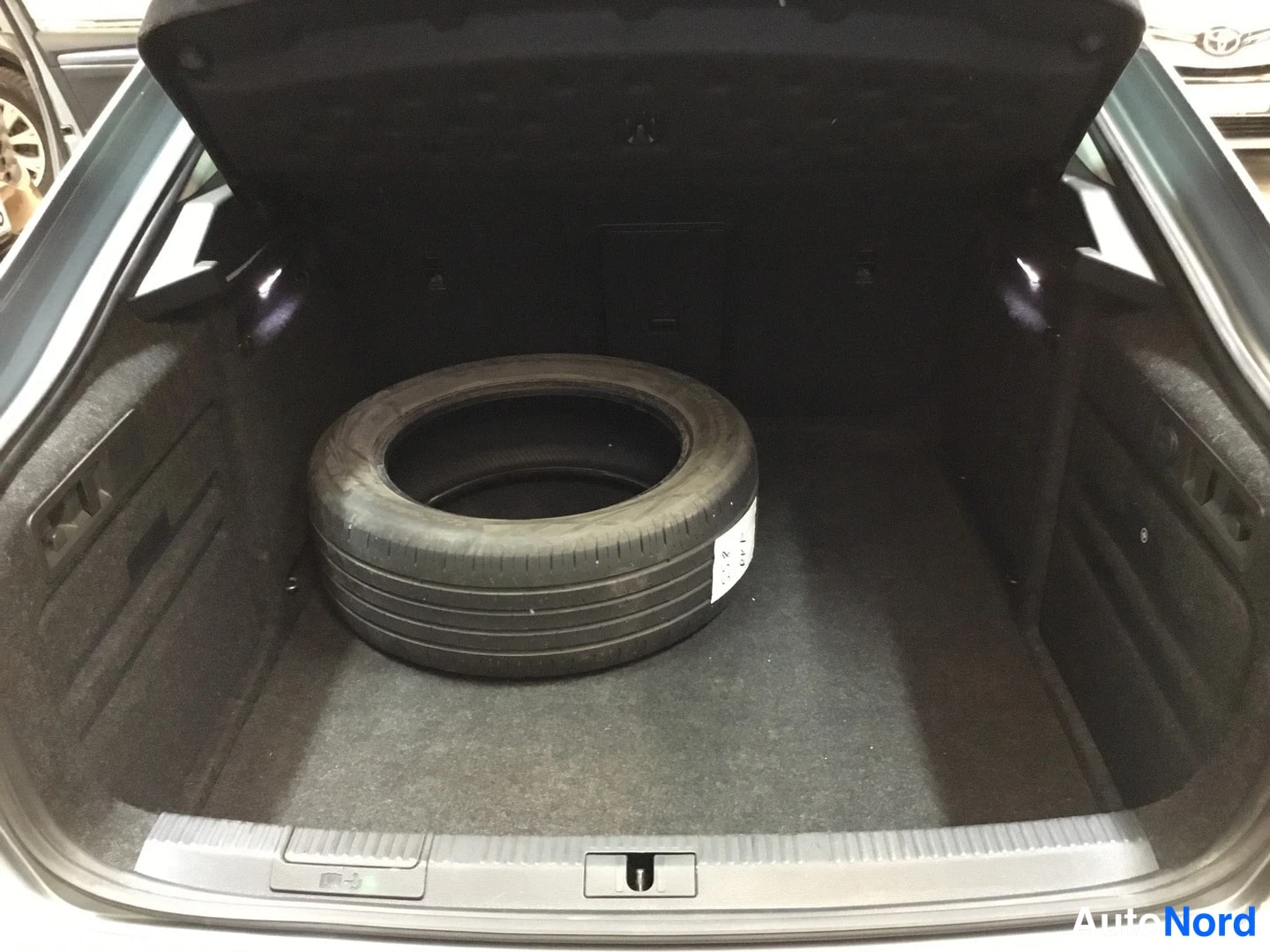 Tavaratila: Open trunk of a car showing a spare tire lying flat on the floor mat. Interior walls are dark grey carpeted with visible tie-down hooks and side panels.