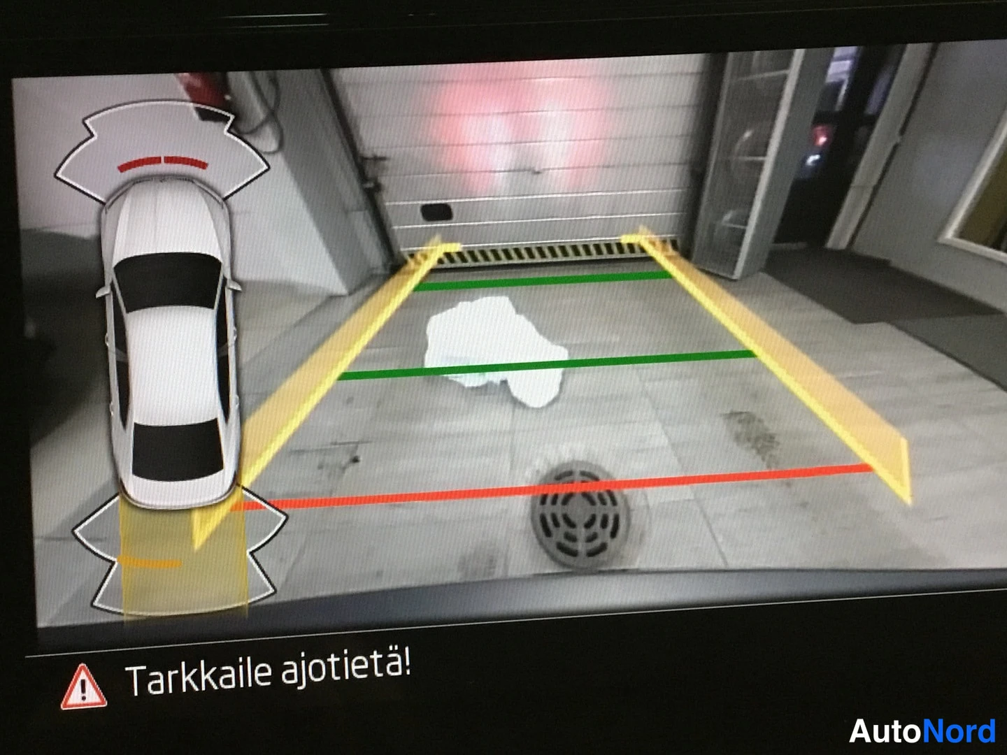 Ohjaamo: Display screen showing a rear view camera image with colored dynamic parking guidelines and a car icon overlay.