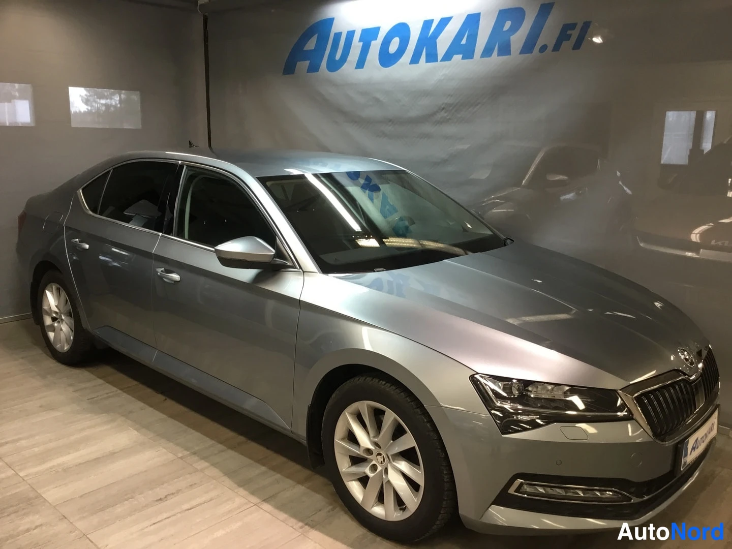 Etuviisto, nokka oikealle: Silver four-door sedan viewed from front-right three-quarter angle in an indoor showroom with tiled floor and a large banner reading AUTOKARI.FI on the w