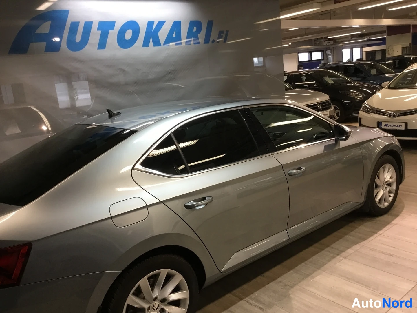 Sivulta: Rear-left side profile of a silver four-door sedan in an indoor showroom, showing rear door, C-pillar, and rear quarter panel.
