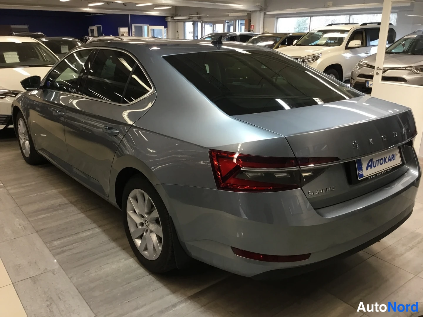 Takaviisto: Rear-right three-quarter view of a silver Skoda Superb sedan in an indoor showroom, showing tailgate, 'SKODA' lettering, and taillights.