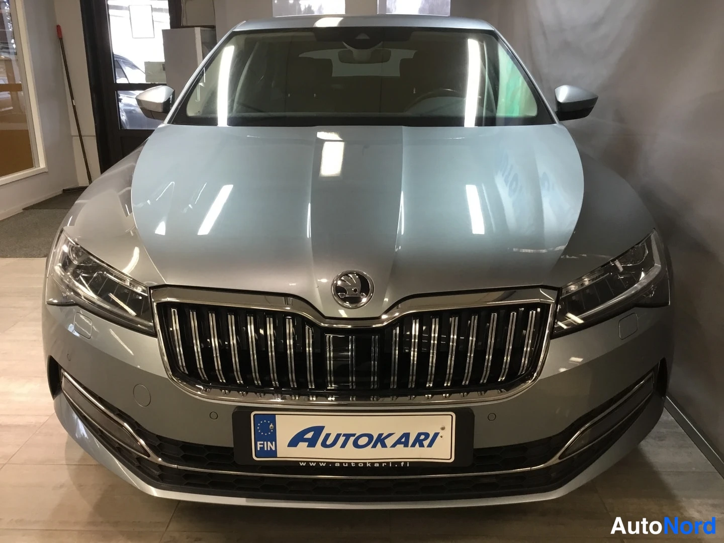 Edestä: Hopeanharmaa Škoda Superb etupäältä, näkyvissä etusäleikkö, ajovalot ja AUTOKARI-etukilpi.