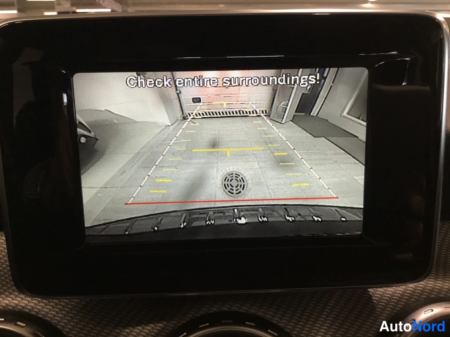 Yksityiskohta: Close-up of the infotainment display screen showing a rear-view camera feed with guidance lines and parking reference graphics. The text 'Check entire surroundi