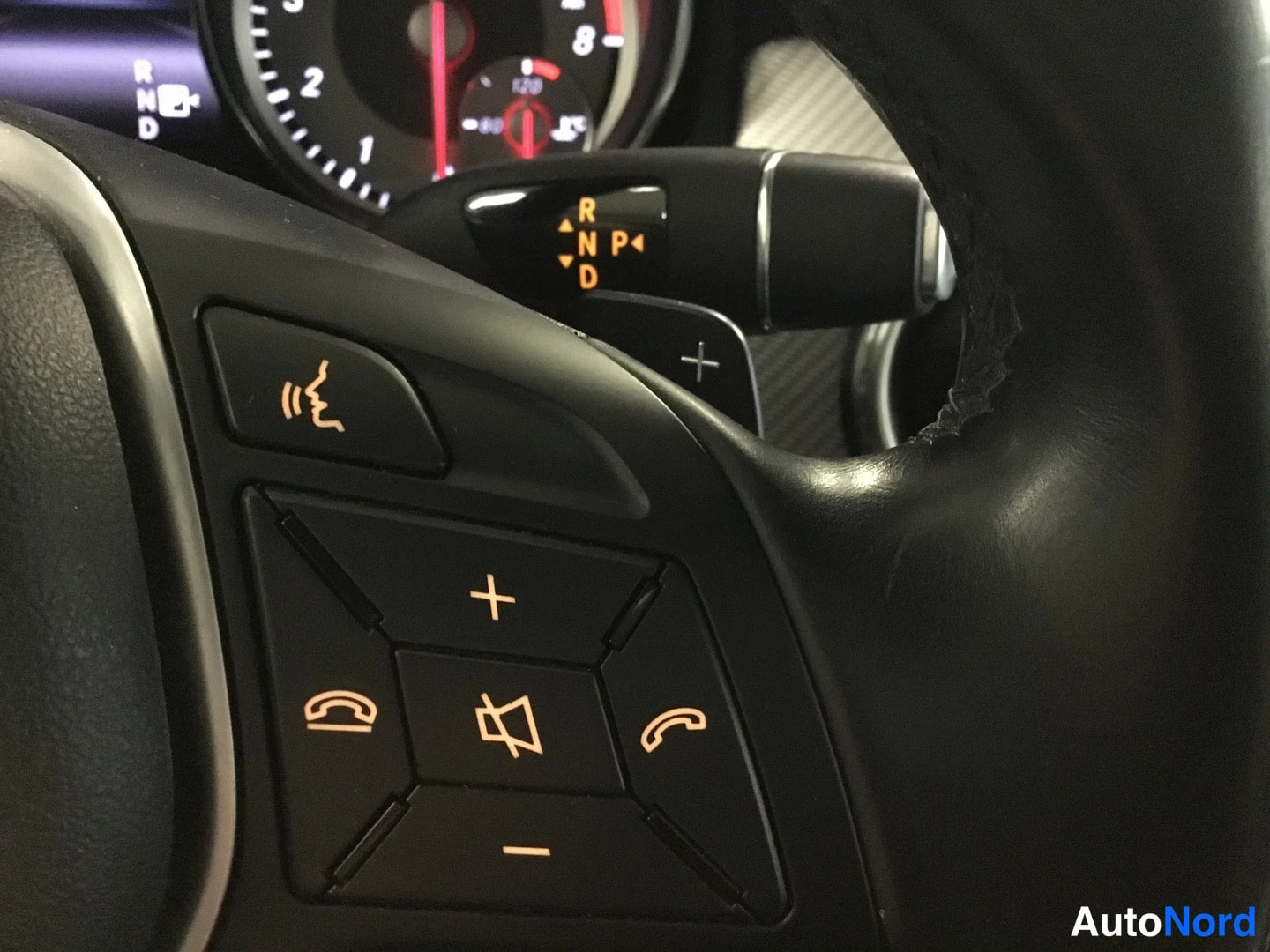 Yksityiskohta: Close-up of the left side of the multifunction steering wheel showing audio controls (volume +/–, track skip) and phone controls (answer/end call). The gear sel