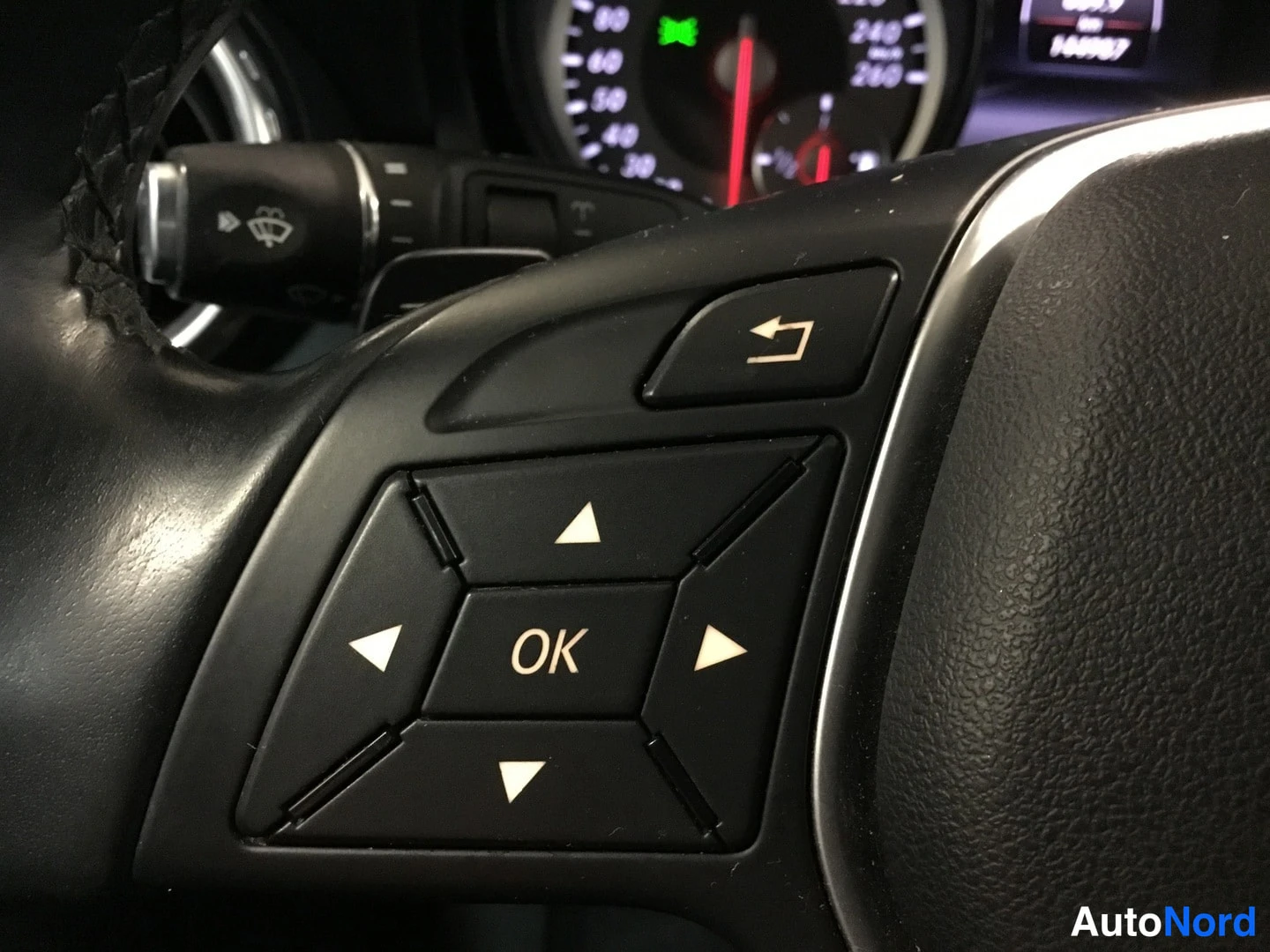 Yksityiskohta: Close-up of the right side of the steering wheel featuring menu navigation buttons (OK, directional arrows, return button). A speedometer with markings up to 26