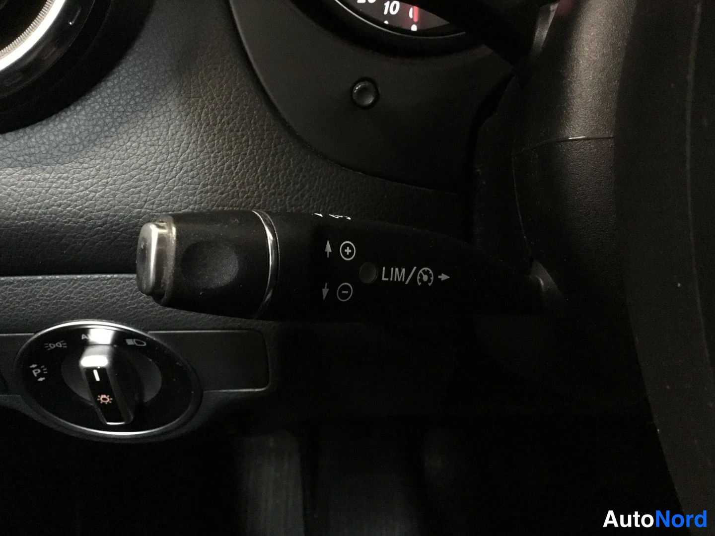 Yksityiskohta: Detailed view of the cruise control stalk mounted on the steering column with '+'/'-' adjusters and 'LIM' limit indicator. Below it is the rotary headlight swit