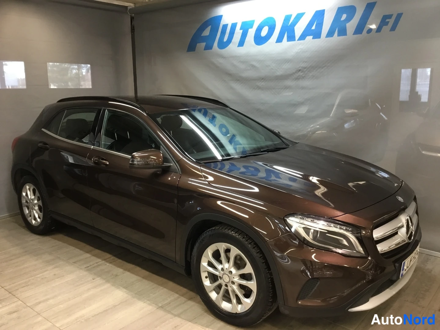 Etuviisto, nokka oikealle: Mercedes-Benz GLA, etukolmikonäkymä vasemmasta sivusta, tummanruskea crossover sisätiloissa Autokari.fi-taustan edessä.