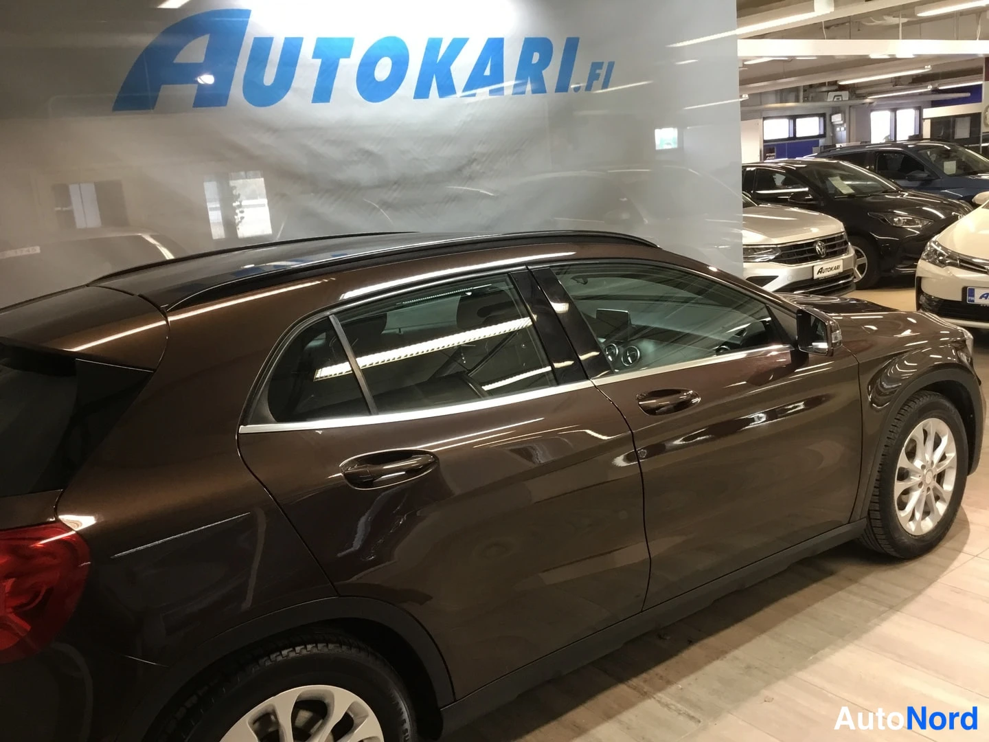 Sivulta: Tummanruskean Mercedes-Benz GLA:n sivunäkymä Autokari.fi-hallissa, takavalaisimen kulku sivun nurkassa.