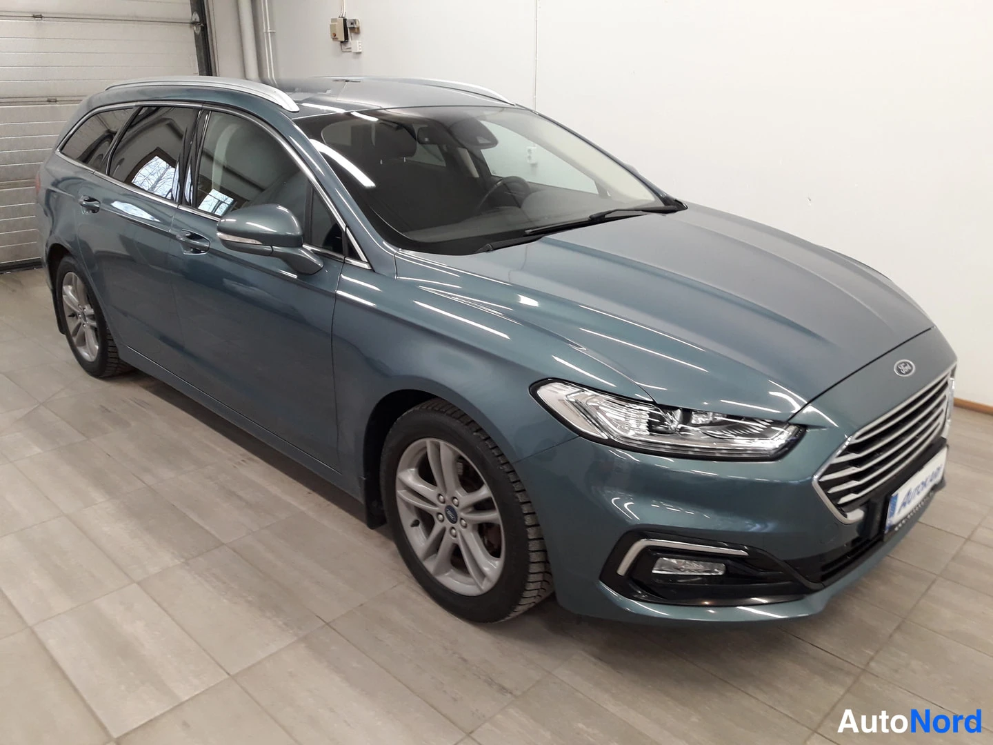 Etuviisto, nokka oikealle: Ford Mondeo wagon in blue-grey color parked indoors on tiled flooring. Front three-quarter angle showing left front corner and driver's side profile wi