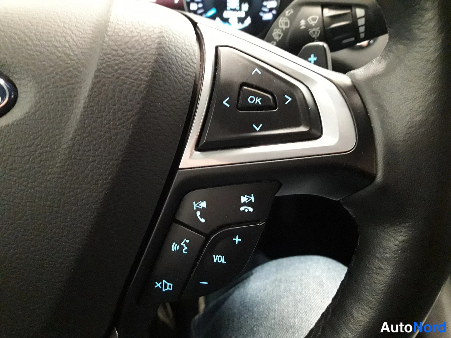 Yksityiskohta: Right side of a black leather steering wheel with volume and media control buttons.