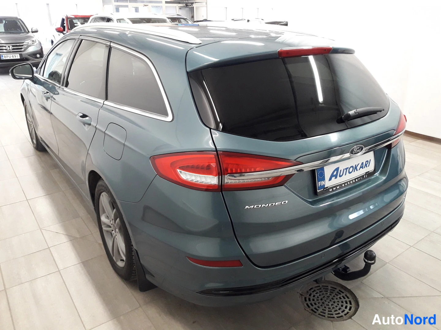 Takaviisto: Rear three-quarter view of Ford Mondeo wagon showing tailgate, taillights with chrome strip, rear bumper with parking sensors and tow hitch. Left rear corner vi