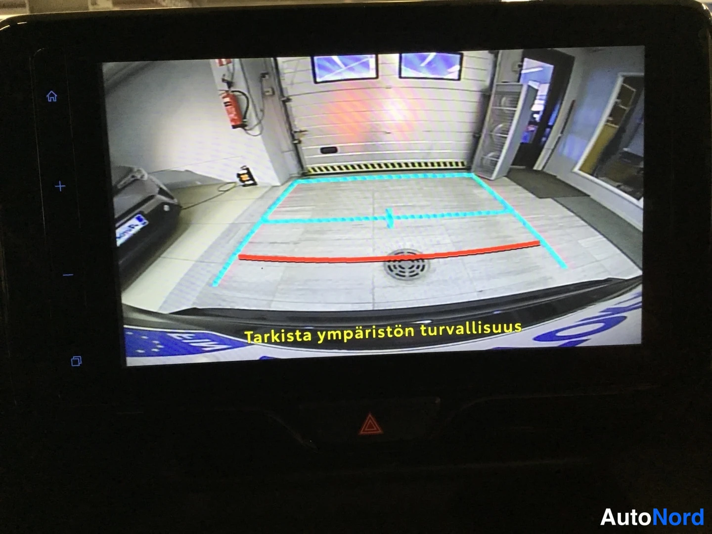 Yksityiskohta: Kamerana näyttö takaa näkymä. Takajarrut ovat näkyvissä ja kameraraidoilla vihreät ja punaiset viivat auttaa ohjausta. Tekstillä suomenkielisellä teksti: 'Tarki