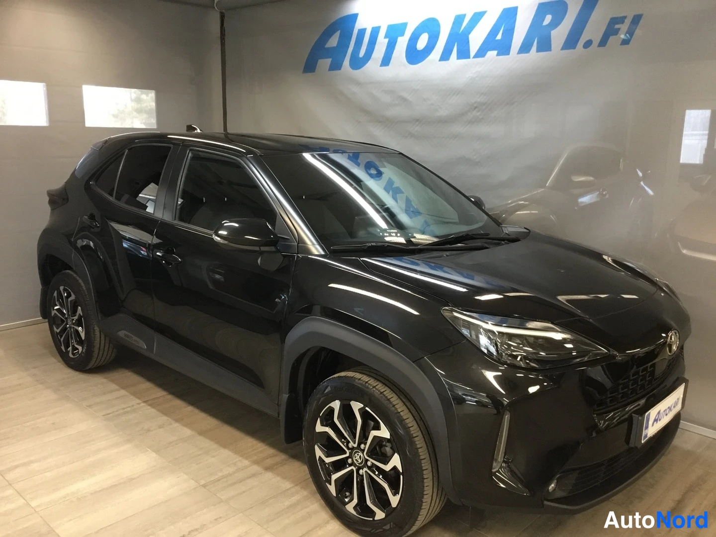 Etuviisto, nokka oikealle: Musta Toyota Yaris Cross -auto edestä nähtynä liikkuvissa kolmesta kulmasta, jossa auton etusivu ja vasen sivu näkyvät; valkoiset rekisterikilven kehät