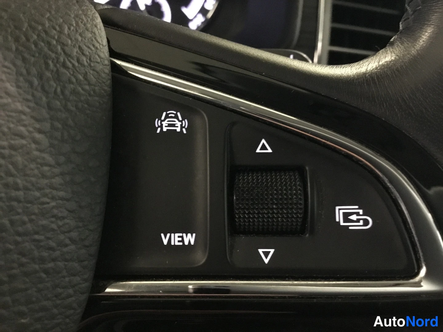 Yksityiskohta: Close-up of the left side of a multifunction steering wheel showing buttons including 'VIEW', directional arrows for display navigation, and an icon representin