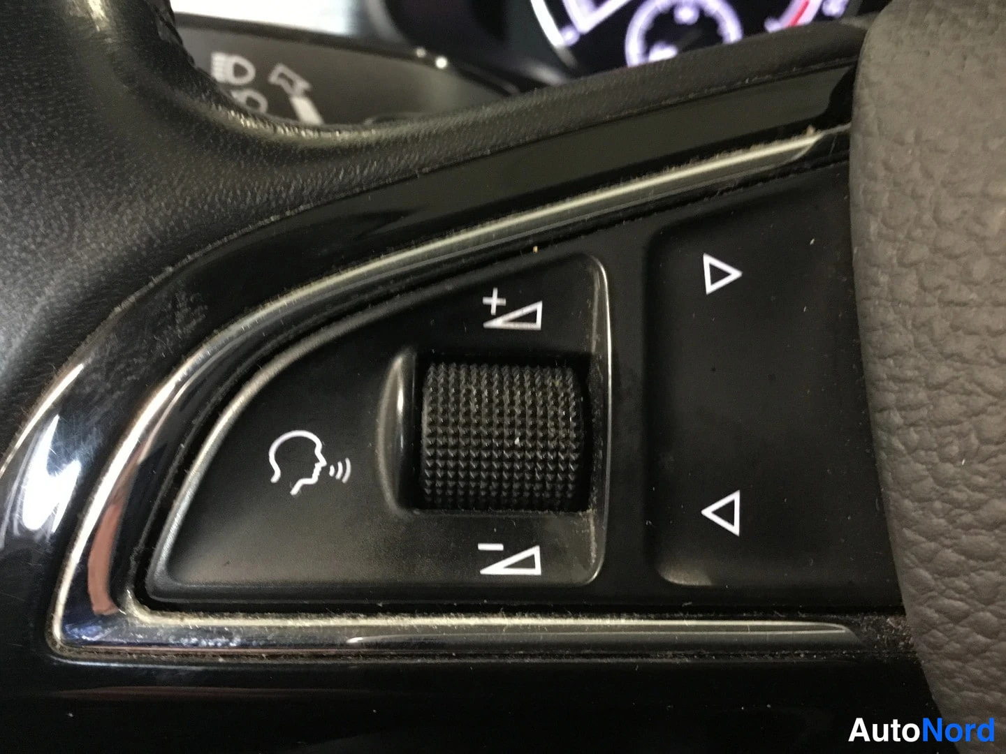 Yksityiskohta: Close-up of the right side of the steering wheel controls featuring volume adjustment scroll wheel, arrow keys for menu navigation, and a voice control button.